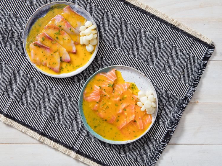 Two shallow bowls of Peruvian tiradito, featuring thinly sliced raw fish drizzled with a bright yellow chili sauce, garnished with herbs and served with Peruvian corn, placed on a patterned woven cloth atop a light wooden surface.