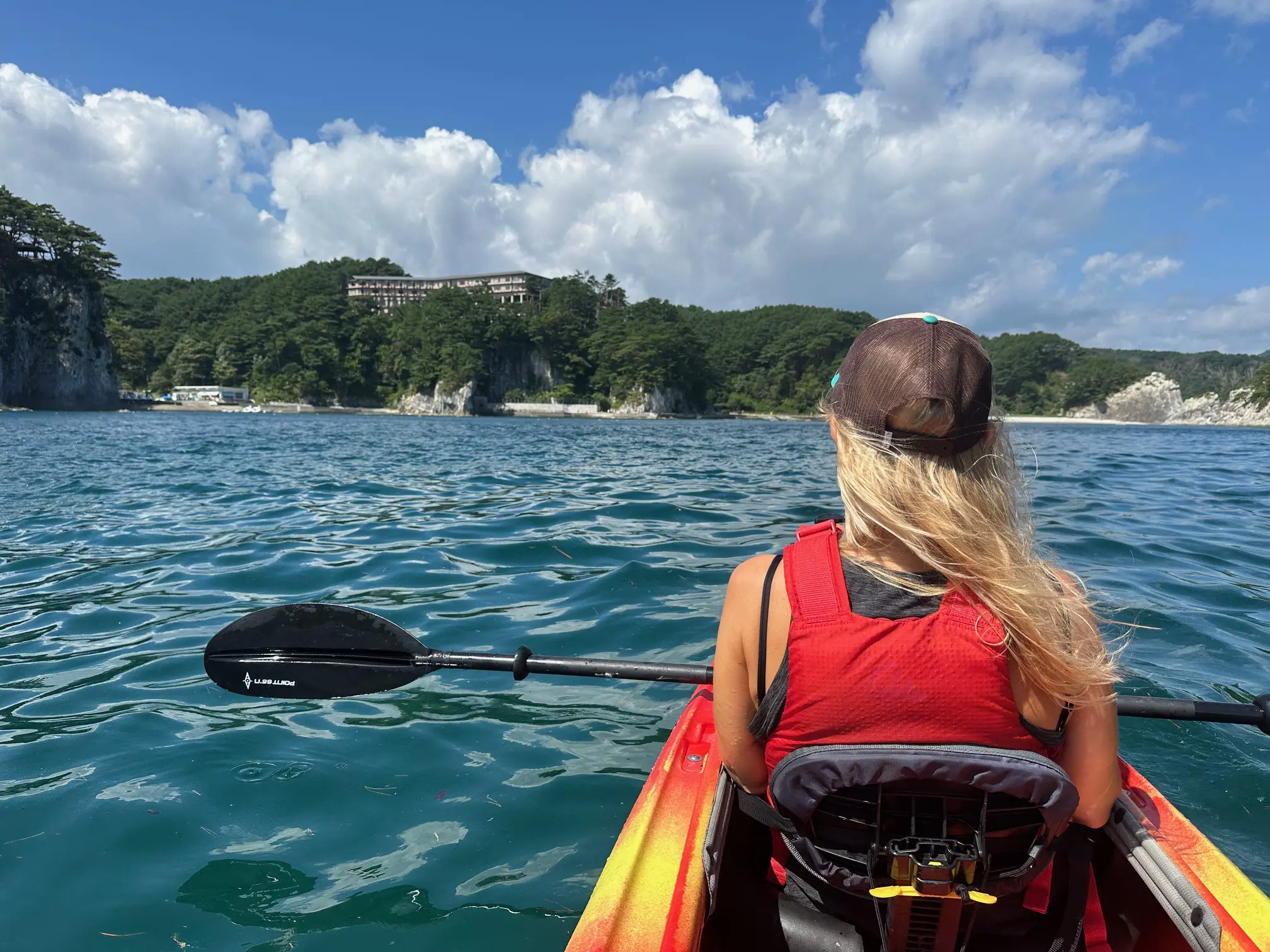 Woman kayaking on the clear blue waters of Jodogahama along the Michinoku Coastal Trail, surrounded by pine-covered cliffs and a bright summer sky.
