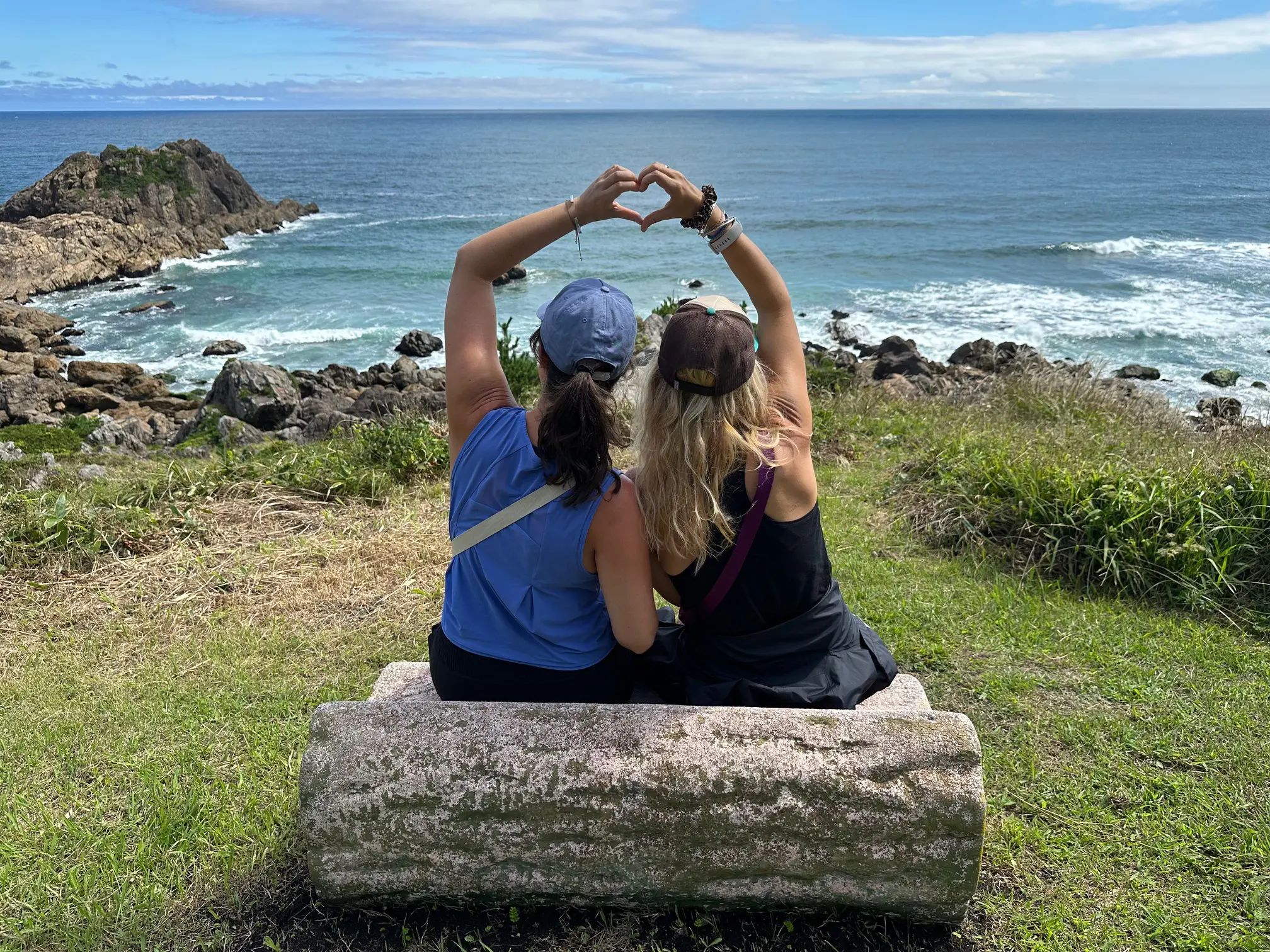Two friends sitting on a bench forming a heart shape with their hands while overlooking the Pacific Ocean along the Michinoku Coastal Trail in Japan