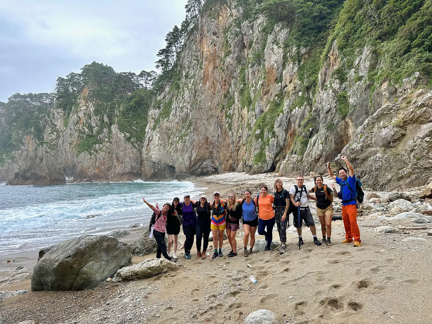 Group of hikers on a beach along the Michinoku Coastal Trail in Japan with rugged cliffs and ocean waves in the background