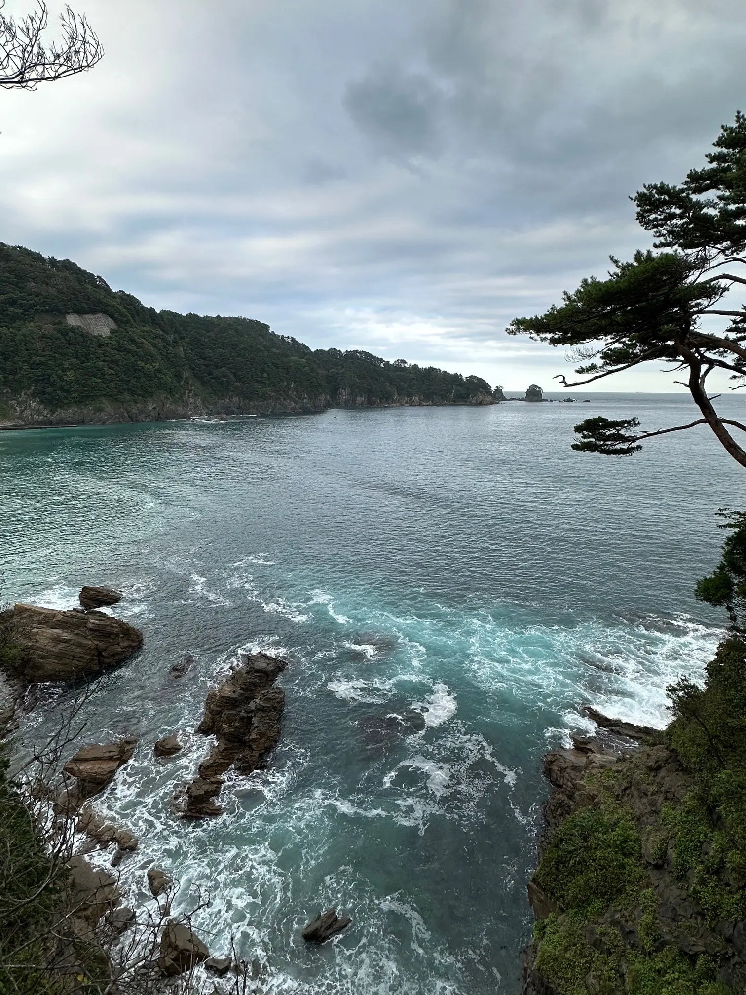 Dramatic coastal view along the Michinoku Coastal Trail in Japan, featuring rocky cliffs, turquoise waves, and forested hills under a cloudy sky